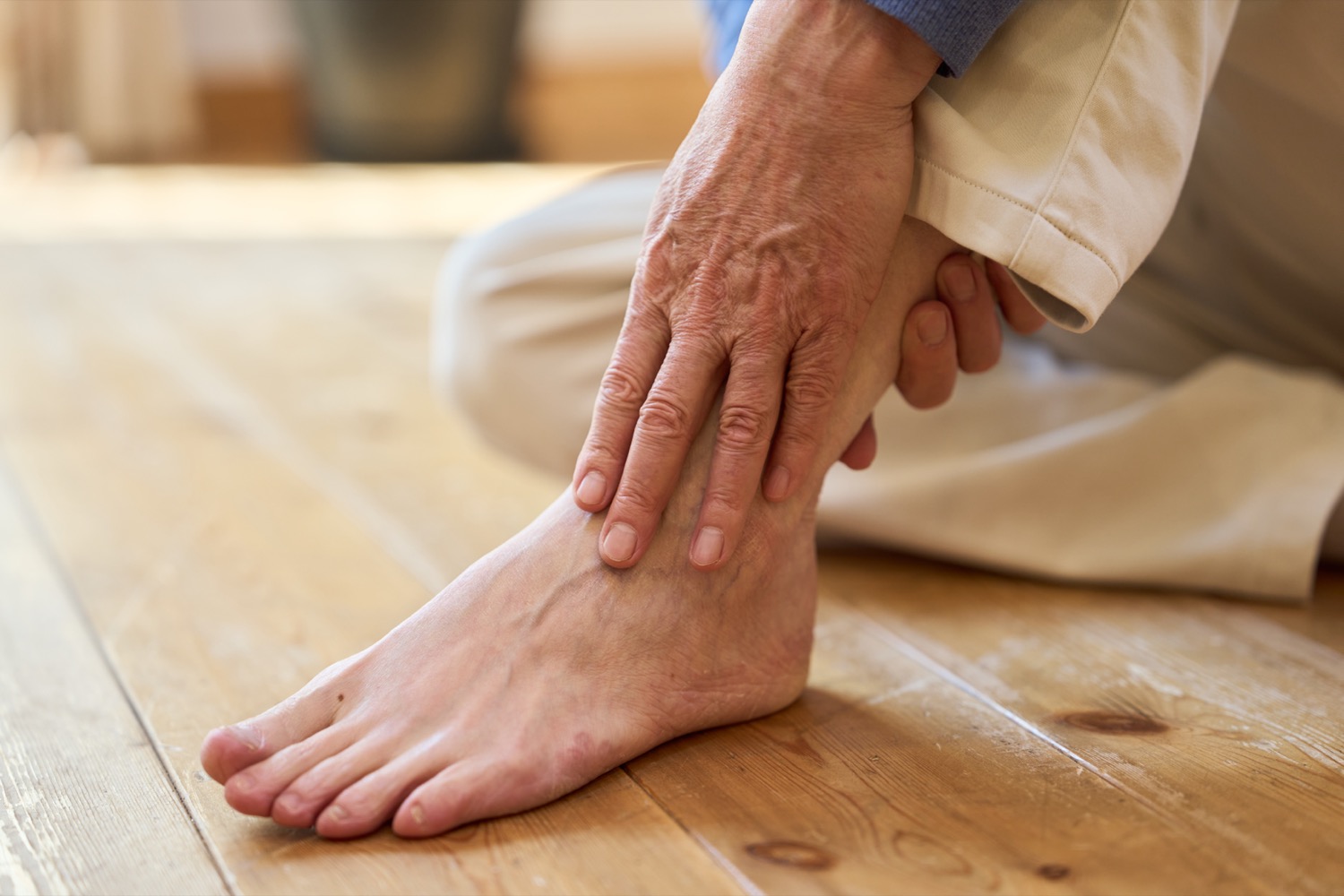 Elderly person holding ankle in pain on wooden floor